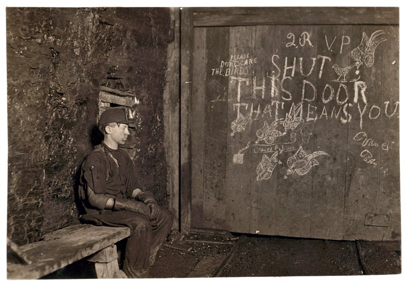 Shut This Door That Means You Vance, a trapper boy, 15 years old. Has trapped for several years in a West Virginia coal mine at 75 cents a day for 10 hours work. All he does is to open and shut this door: most of the time he sits here idle, waiting for the cars to come. On account of the intense darkness in the mine, the hieroglyphics on the door were not visible until plate was developed. September 1908. View full size. Photograph and caption by Lewis Wickes Hine.
One trapper's description of the job, which paid about $1.60 a day:
Trappers were responsible for opening and closing the underground ventilation doors. In those old mines, they had a system of doors between sections to direct the flow of air. Air was supposed to go up the main haulage and back to the fan. So a trapper sat all day by his door with an oil lamp on his cap. There was a "manhole" - a shelter hole in the wall by the track. The motorman would blink his light at me, and I'd throw the switch and open the door for him. Then, I'd jump into the manway until he was past, and run out and close the door. A trip would come along about every hour. Was I bored or lonely? Well, it was my job.