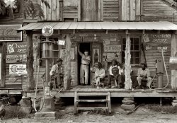 July 1939. Gordonton, N.C. "Country store on dirt road. Sunday afternoon. Note kerosene pump on the right and the gasoline pump on the left. Rough, unfinished timber posts have been used as supports for porch roof. Negro men sitting on the porch. Brother of store owner stands in doorway." 4x5 nitrate negative by Dorothea Lange for the Farm Security Admin. View full size.